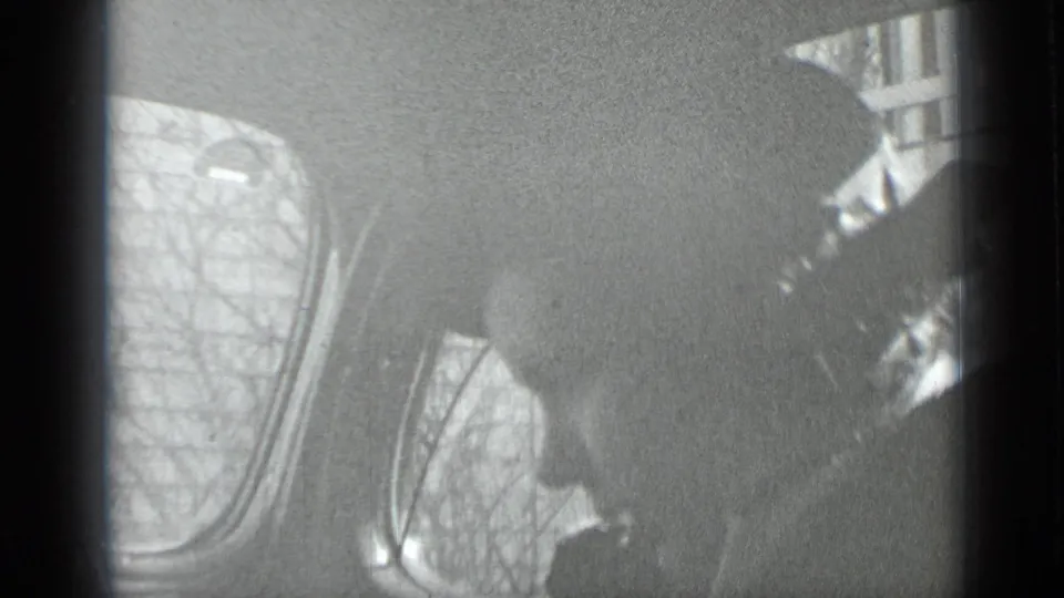 Black-and-white 1939 home-movie frame from New York showing a shadowed person inside a vehicle, with rounded windows and leafless trees beyond.