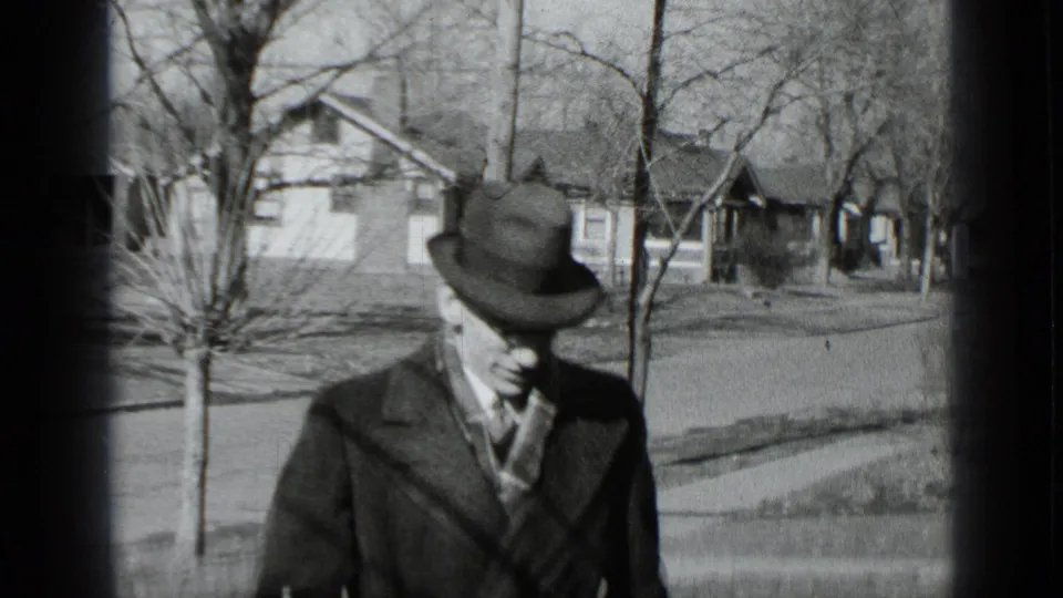 Black-and-white 1939 New York home-movie frame showing a man in a fedora and dark overcoat walking through a quiet residential street.