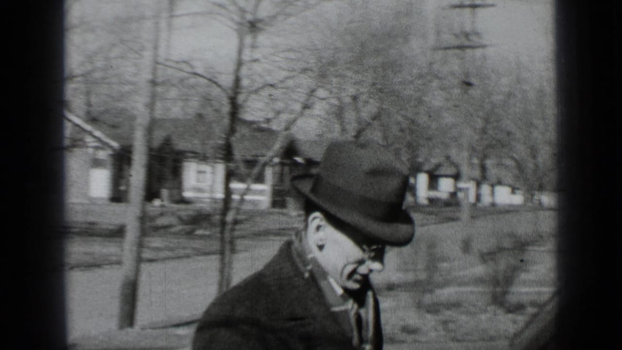 Man in fedora and coat walks on a residential street with bare trees and houses behind him.