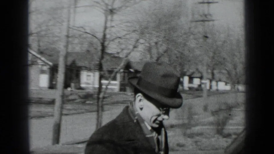 In 1939 New York home-movie footage, a man in a fedora and dark coat walks past modest houses on a quiet street.