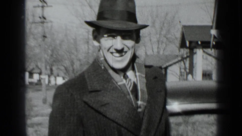 A smiling man in a fedora and overcoat looks toward the camera on a New York residential street in 1939.