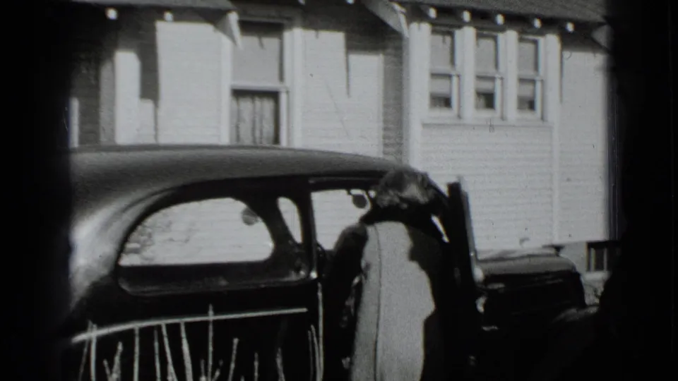 A person stands by the open door of a dark period car outside a residential house in black-and-white 8mm footage.