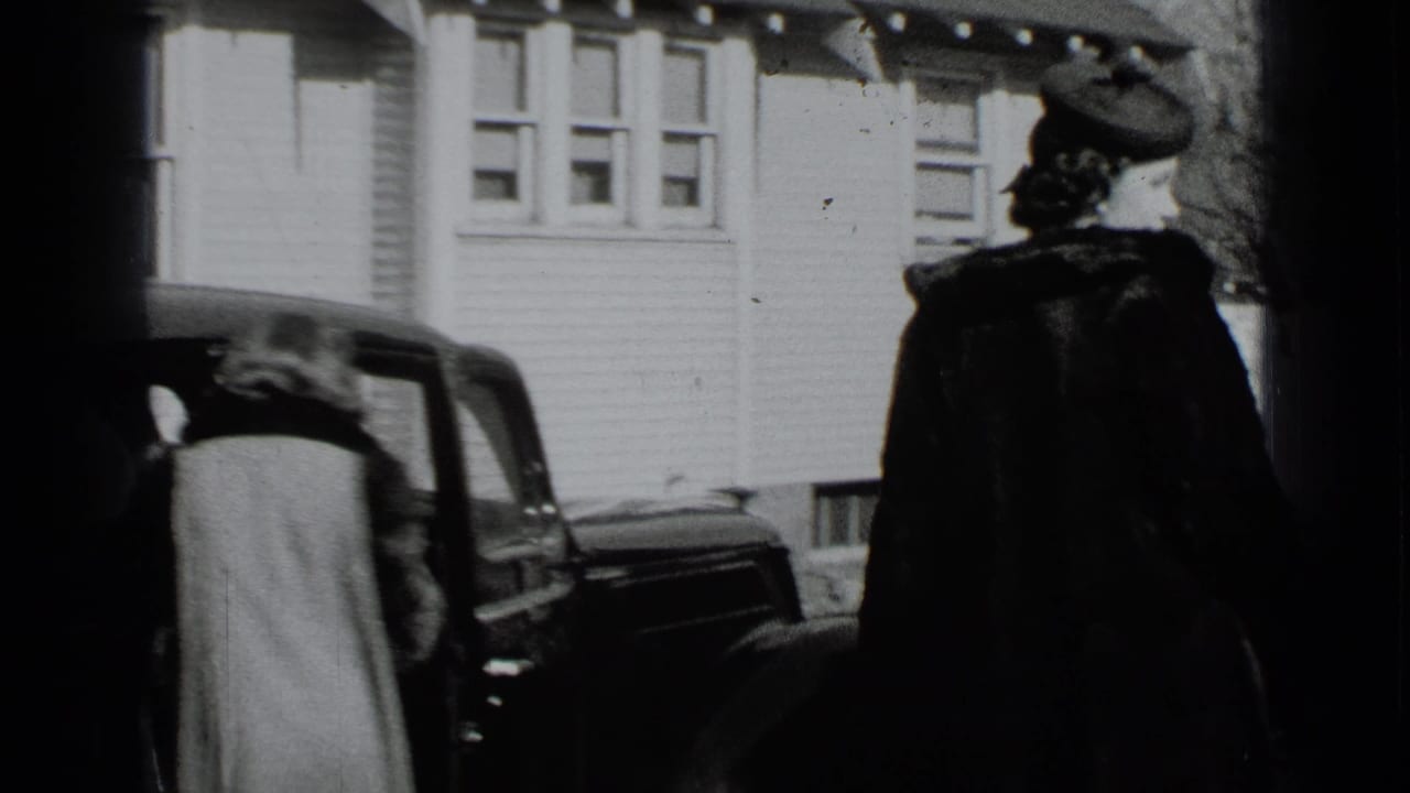 Two women stand beside a dark vintage car in front of a white clapboard house.