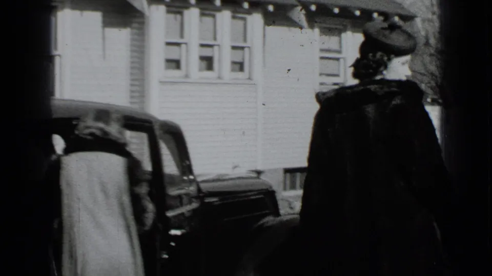 In 1939 New York home-movie footage, two women in coats stand near a dark car outside a white clapboard house.