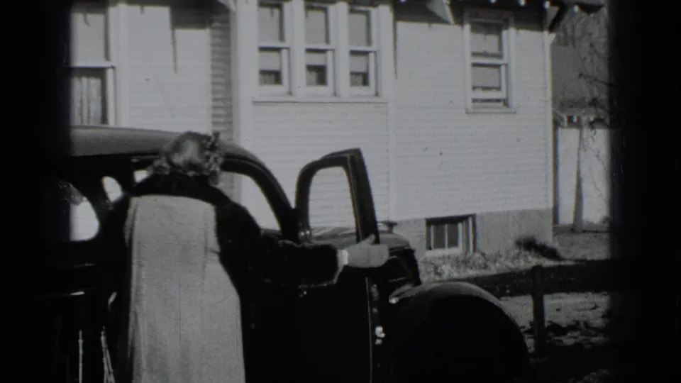 A woman in a light coat stands beside an open dark car door outside a white clapboard house in 1939 New York footage.