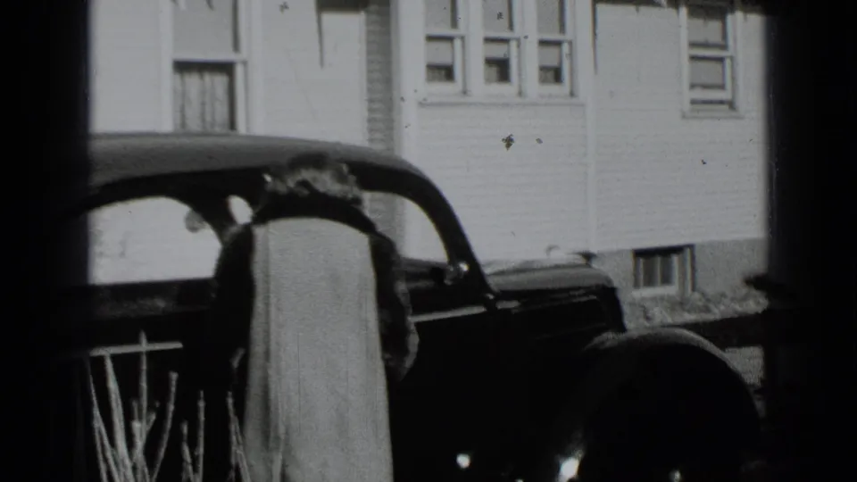 The woman remains beside the dark automobile outside the clapboard house as the brief 1939 New York home-movie scene ends.