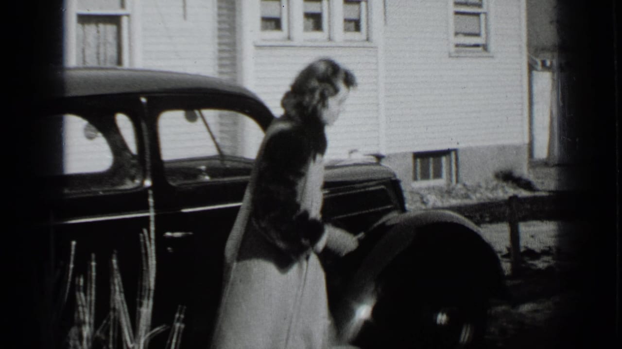Woman beside a dark car in front of a clapboard house.