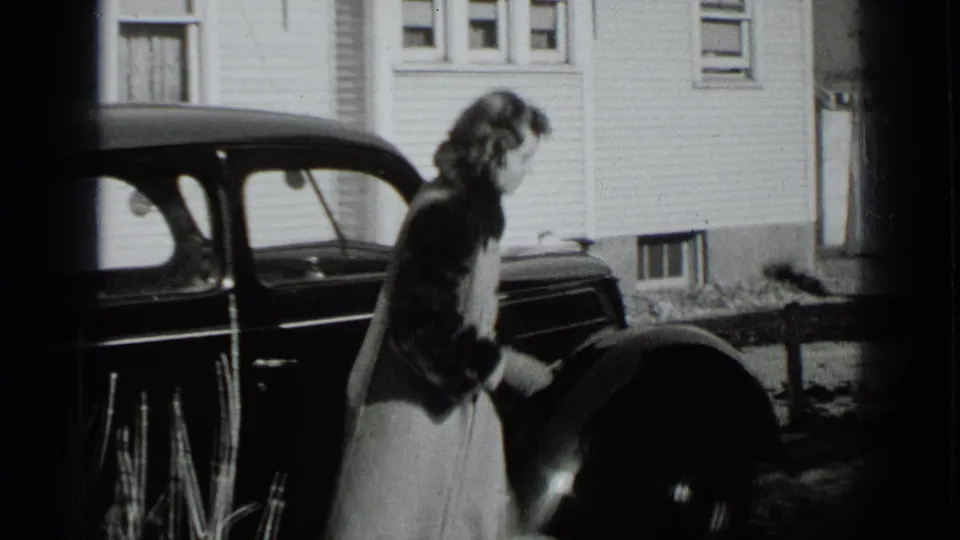 In 1939 New York home-movie footage, a woman in a long coat stands beside a dark car outside a clapboard house.