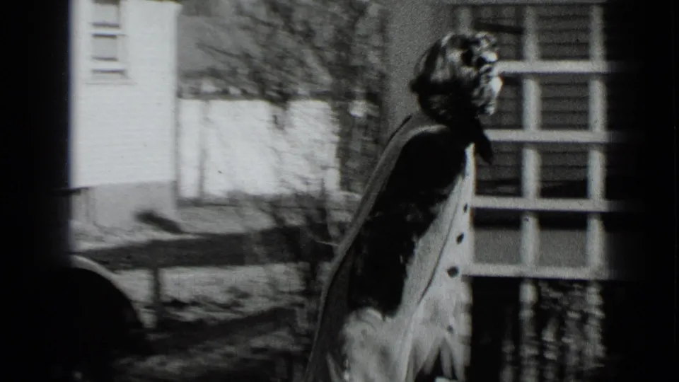 The woman walks from the parked car toward the house exterior, her coat and hairstyle visible in profile.