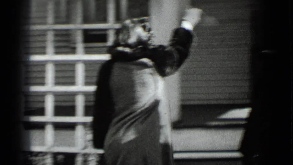 Near the clapboard house, the woman raises one arm as if tossing or signaling toward the doorway.