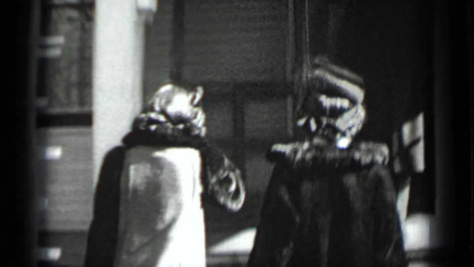 The woman reaches the entrance area where another adult stands near the doorway of the house.