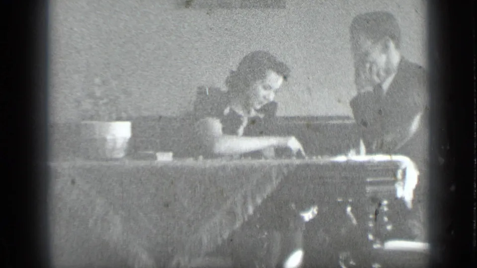 Inside, a man and woman sit at a table, possibly handling tabletop items during a quiet home-movie moment.