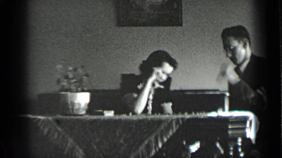 A woman leans on her hand at the chess table while the man shifts position in the grainy 8mm interior scene.