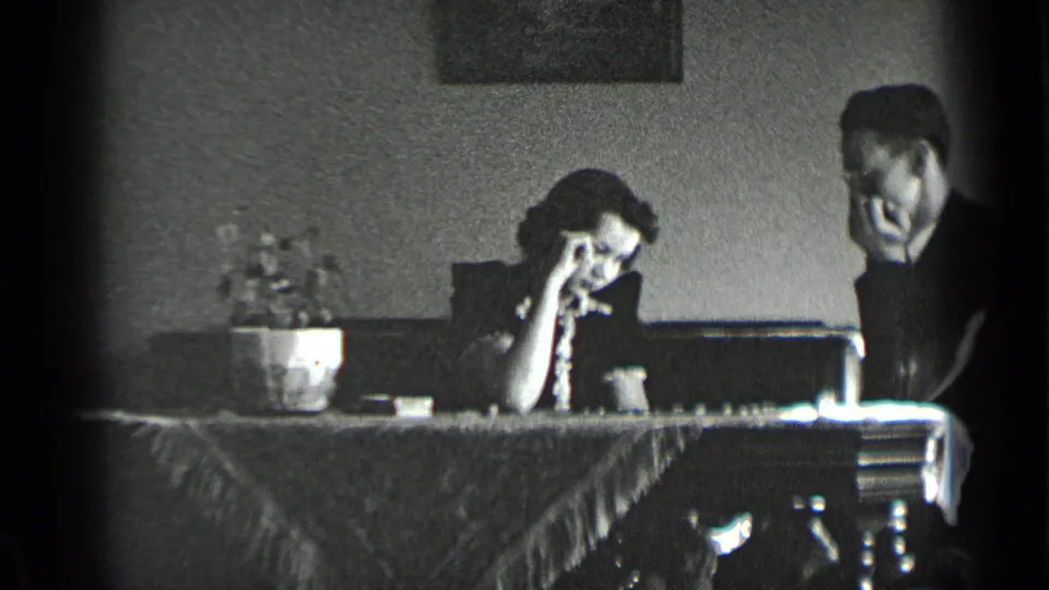 The couple remains seated at the chess table, with the woman looking down and the man beside her in a private home interior.