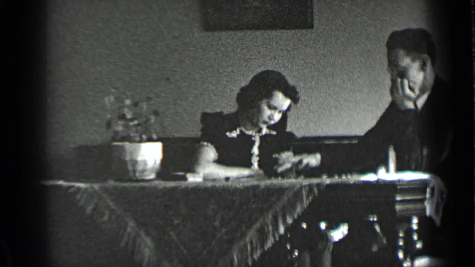 In a 1939 New York home-movie frame, a woman looks down at a tabletop activity while a man sits beside her with his hand at his face.
