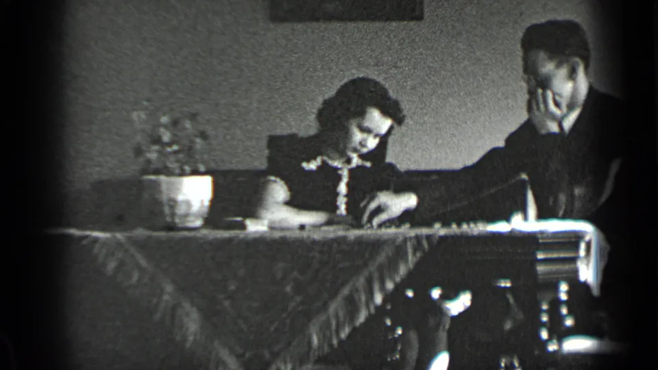The 1939 New York interior frame shows the woman focused downward as the man extends a hand toward small objects on the covered table.
