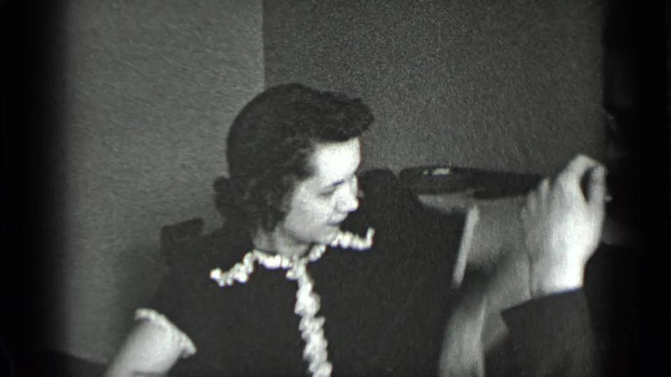 A closer 1939 New York frame shows the woman in a dark dress with a light ruffled collar turning toward a raised hand near the table.