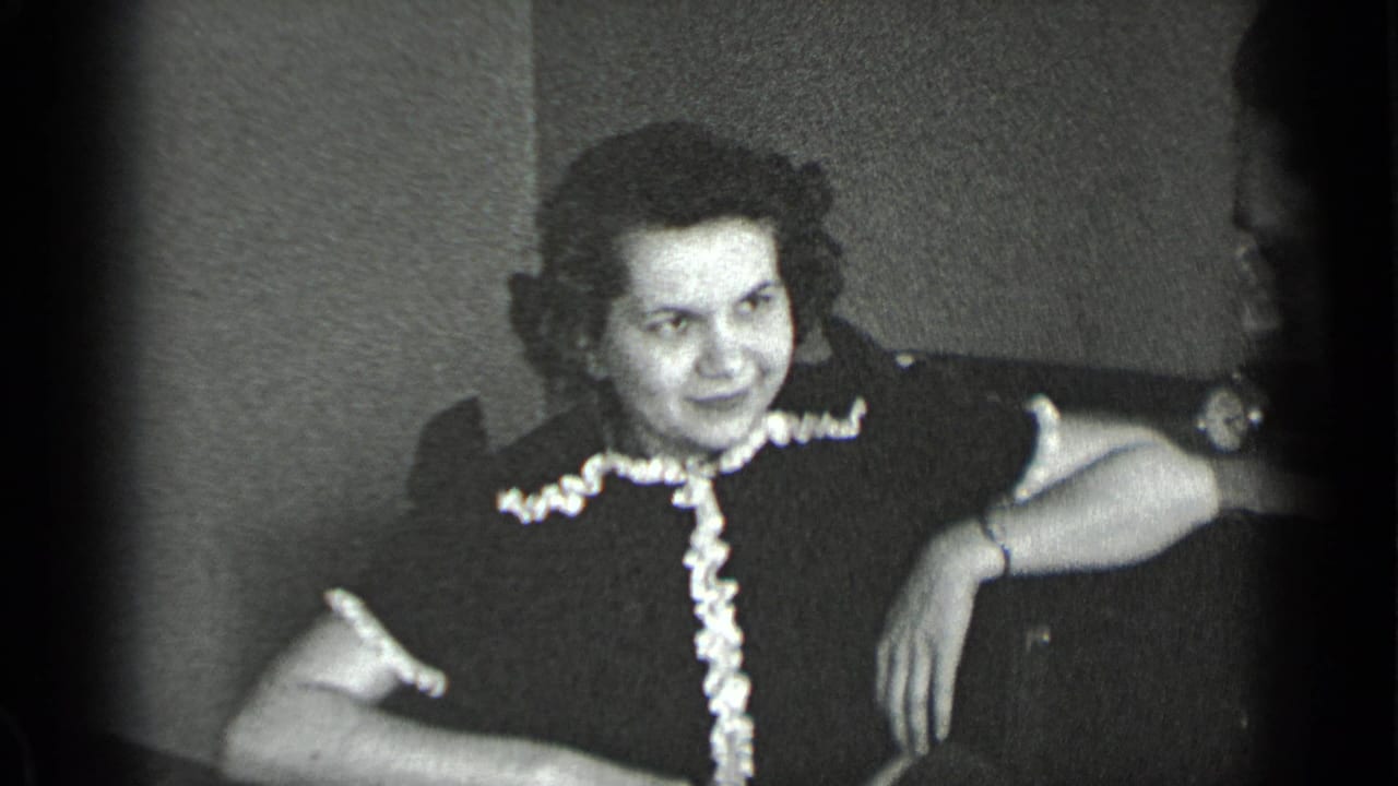 Woman seated indoors in a dark dress with light ruffle trim, smiling beside furniture.