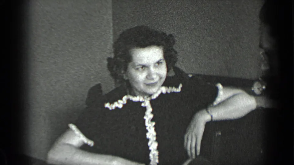 1939 New York home-movie frame of a seated woman indoors, wearing a dark dress with light ruffle trim and smiling toward someone nearby.