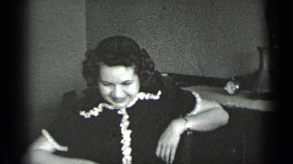 Seated woman in a dark ruffled dress looks down inside a home interior, with furniture and film edge shadows visible.
