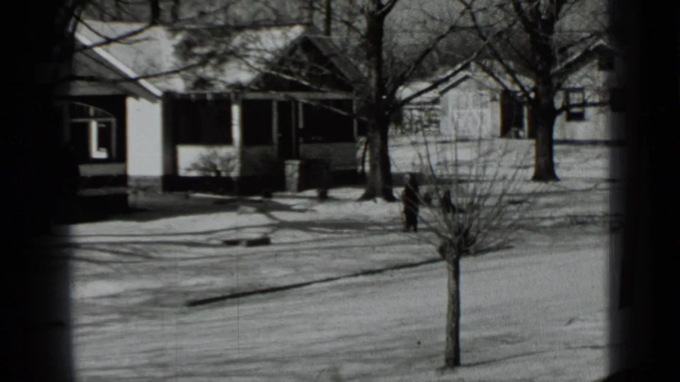Snowy New York residential exterior from 1939 home-movie footage, showing houses, bare trees, a street, and a distant person.