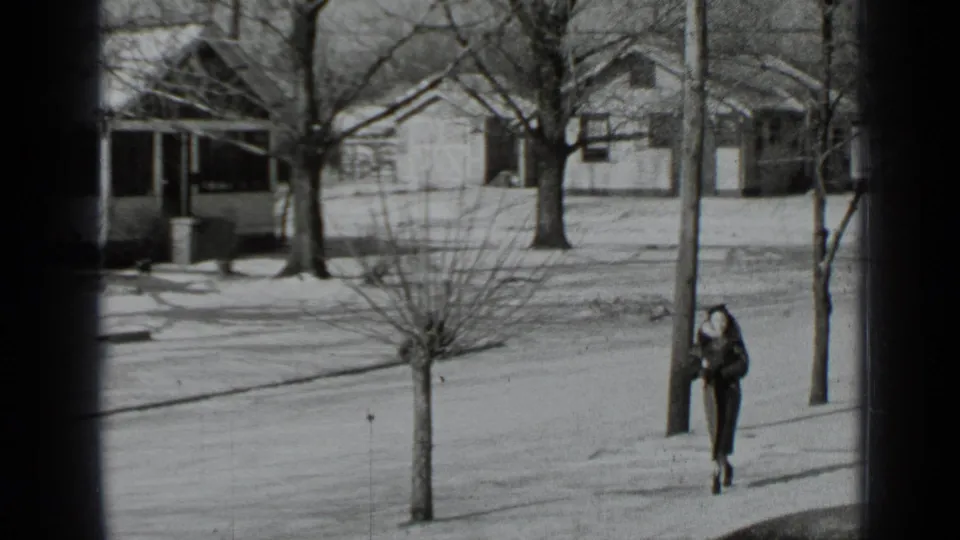 In 1939 New York home-movie footage, a woman in a long coat walks along a snow-dusted residential sidewalk.
