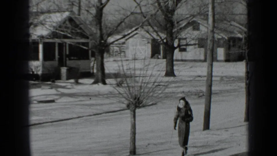 The woman continues down a snow-dusted New York residential sidewalk, with bare trees and houses in the background.