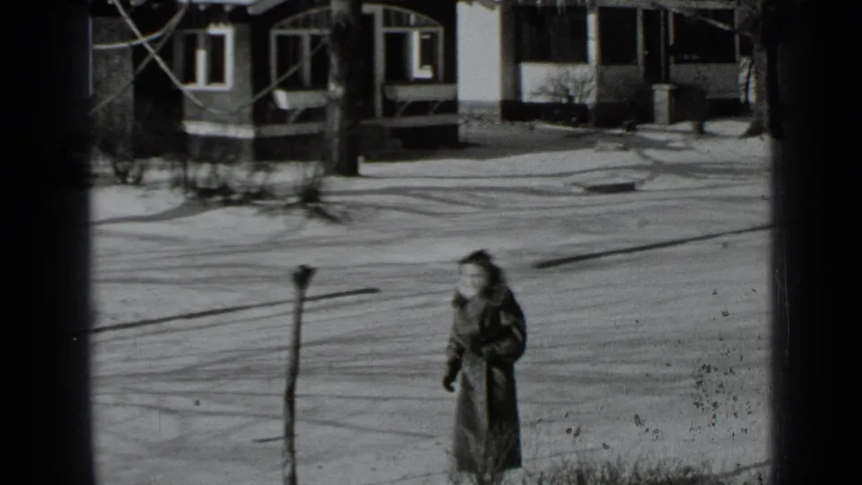 The woman continues down the snow-dusted sidewalk, framed by bare trees and modest houses in 1939 New York.