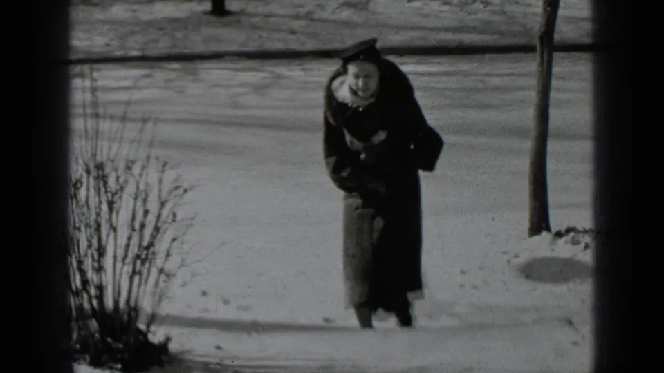 A closer view shows the bundled woman approaching the camera on a snowy sidewalk in a quiet New York neighborhood.