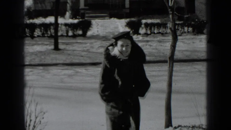 In 1939 New York home-movie footage, a woman in a dark winter coat and hat walks on a snowy sidewalk near bare trees.