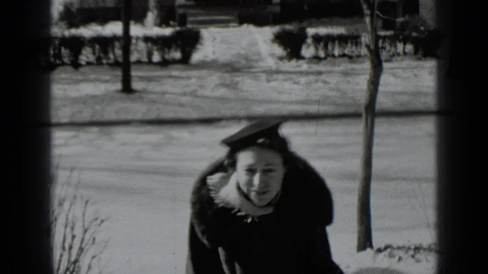 The woman in a heavy coat and hat looks toward the camera on a snowy New York sidewalk, with hedges and bare trees behind her.