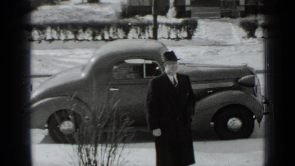 A man in a dark overcoat and brimmed hat stands on a snowy street beside a rounded 1930s automobile in New York footage.