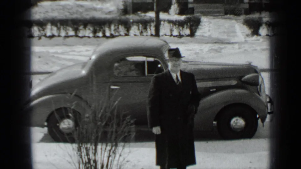 The man remains beside the parked 1930s automobile, facing the camera on a snowy New York residential street.