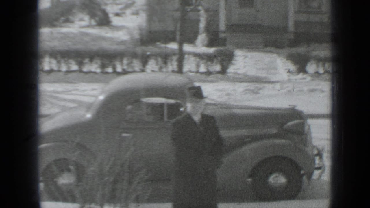 In 1939 New York home-movie footage, a man in a long coat and hat stands next to a dark automobile on a snow-covered residential street.