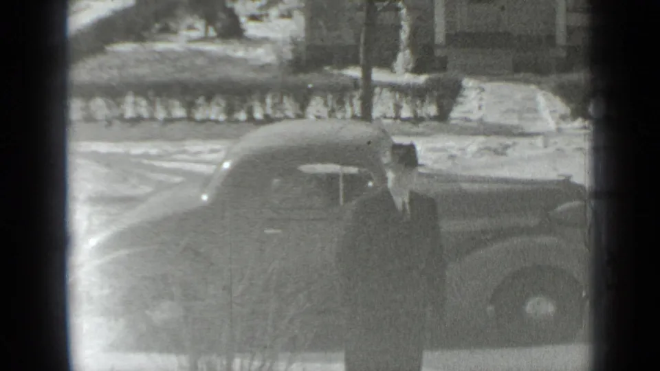 A dark 1930s automobile sits along a snow-lined residential street in New York, with houses, hedges, and bare winter trees visible behind it.