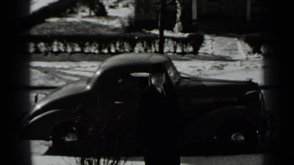 A man in a dark coat and brimmed hat stands near the side of a 1930s car, with snow and a residential sidewalk behind him.
