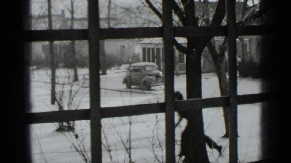 1939 New York home-movie frame showing a woman skating in a snowy residential yard, viewed through a window with dark mullions.