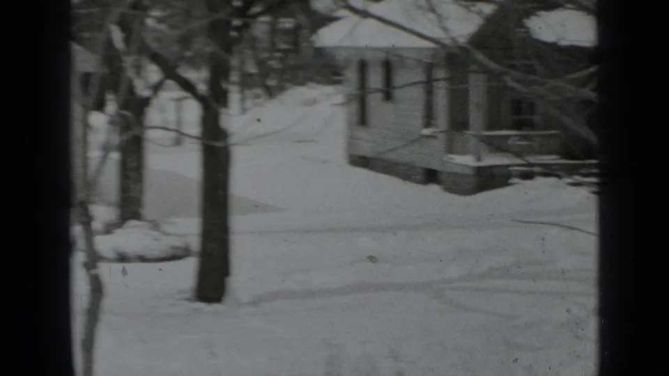 1939 New York home-movie frame of a snowy residential yard and house, partially blocked by dark window mullions and tree trunks.