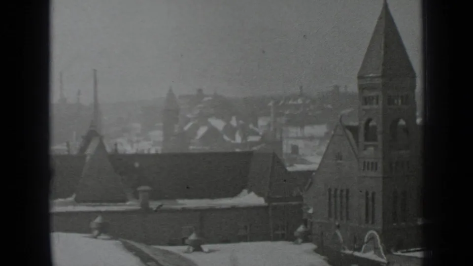 Grainy black-and-white 1939 New York view of a church tower, snow-covered roofs, chimneys, and distant urban buildings through glass.