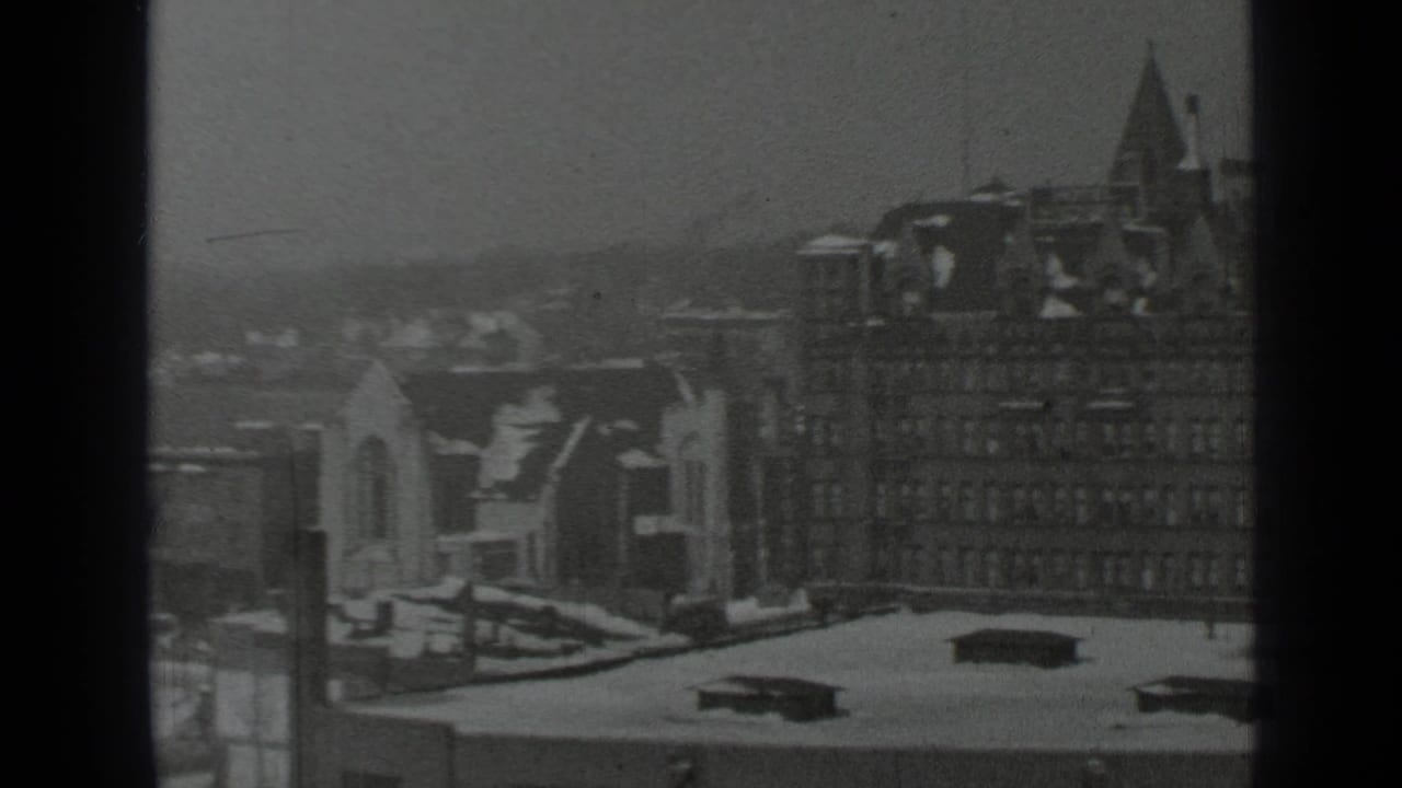 Snowy city rooftops seen through dark window mullions with a large masonry building at left.