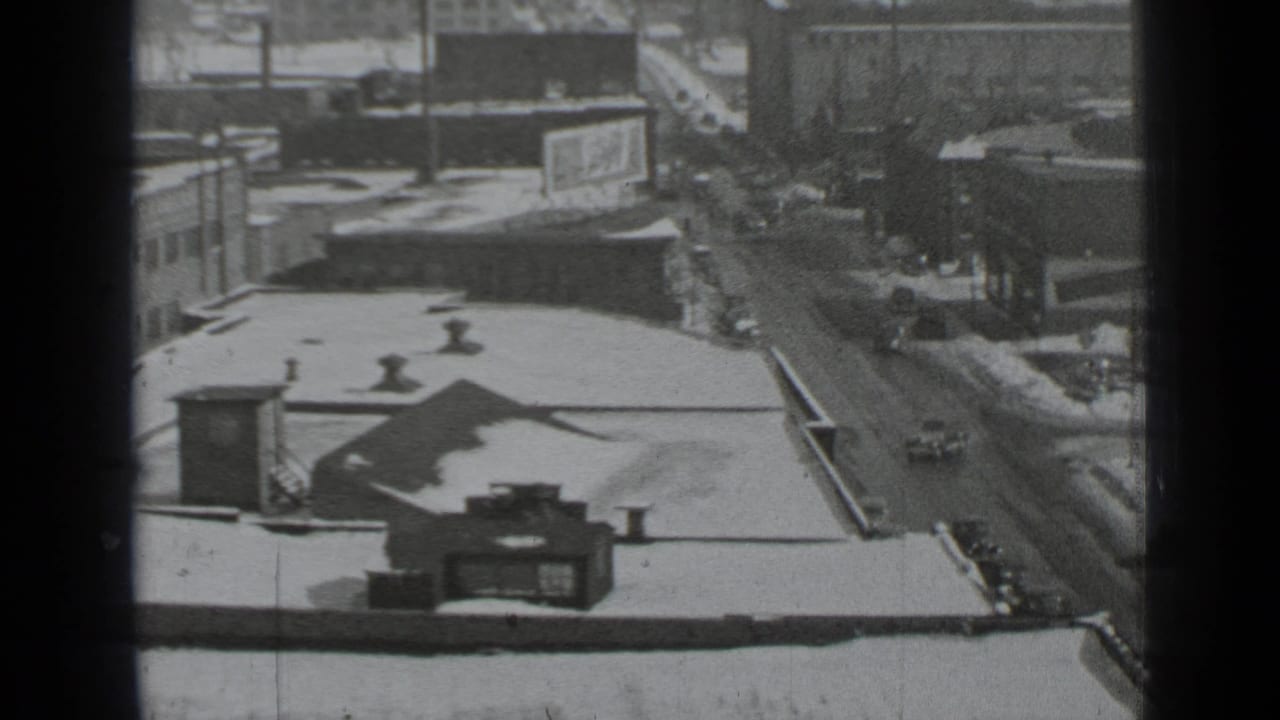 Snow-covered rooftops and wet city street seen from above with cars below.