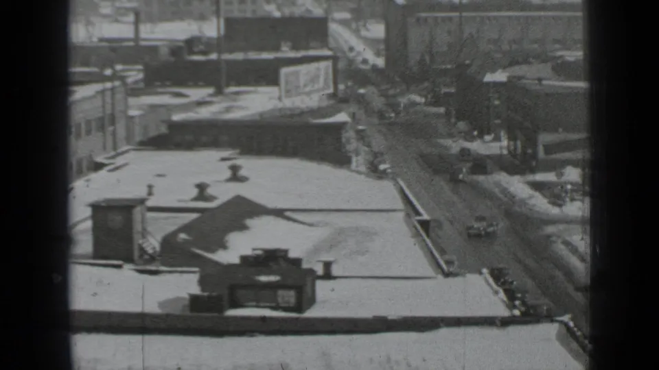 High-angle 1939 New York street view with snowy rooftops, wet pavement, vintage cars, and dense mid-rise buildings.