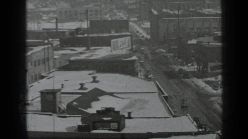 Elevated black-and-white view of snowy New York rooftops, a wet traffic corridor, a billboard, and distant buildings.