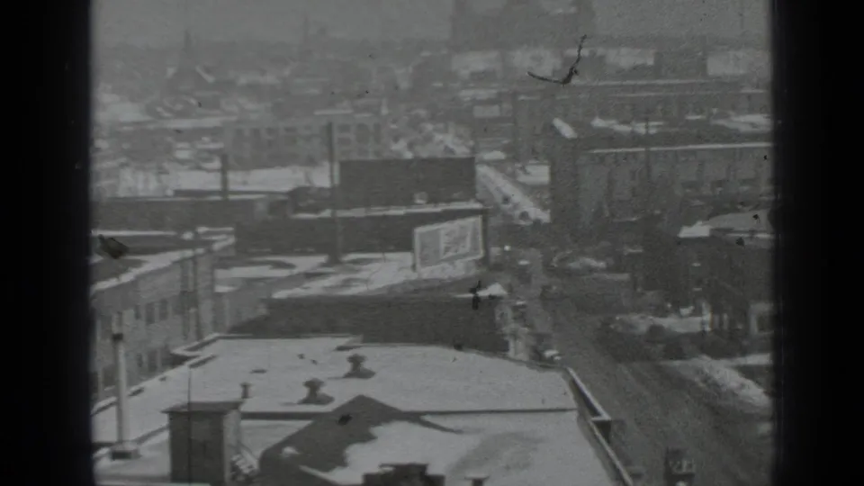 Grainy 8mm frame of a snowy New York city block, wet roadway, older cars, billboard, and hazy background buildings.