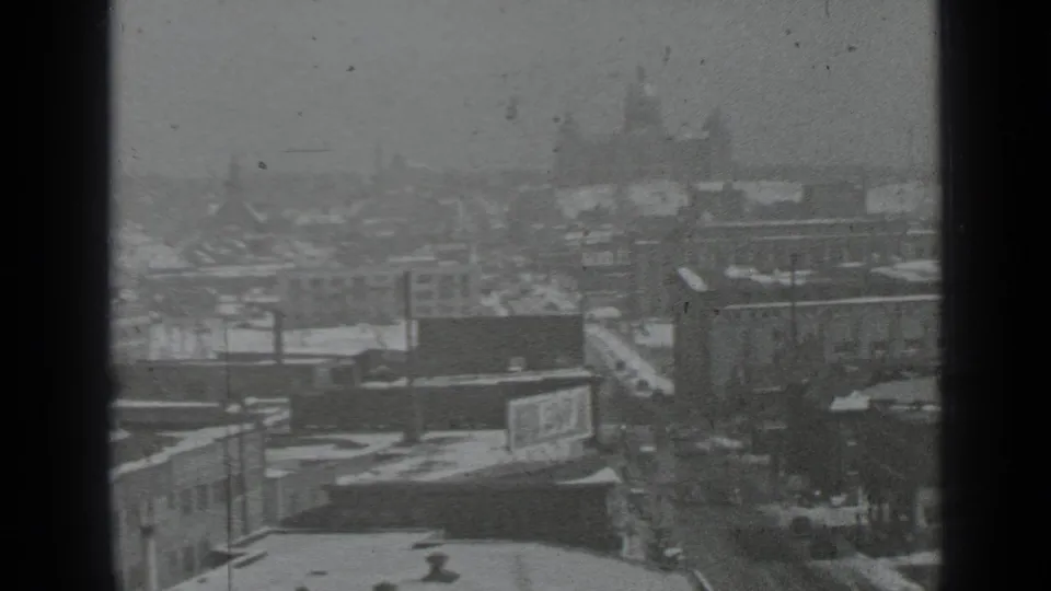 Elevated 1939 New York winter view with snow-covered rooftops, wet street traffic, billboard, and a hazy domed building in the distance.