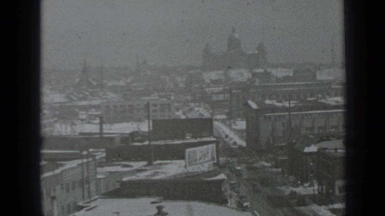 Snowy urban view through dark window frames with streets, rooftops, and distant buildings.