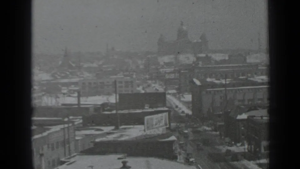 1939 New York snowy cityscape seen from above through window frames, with rooftops, streets, and distant mid-rise buildings.