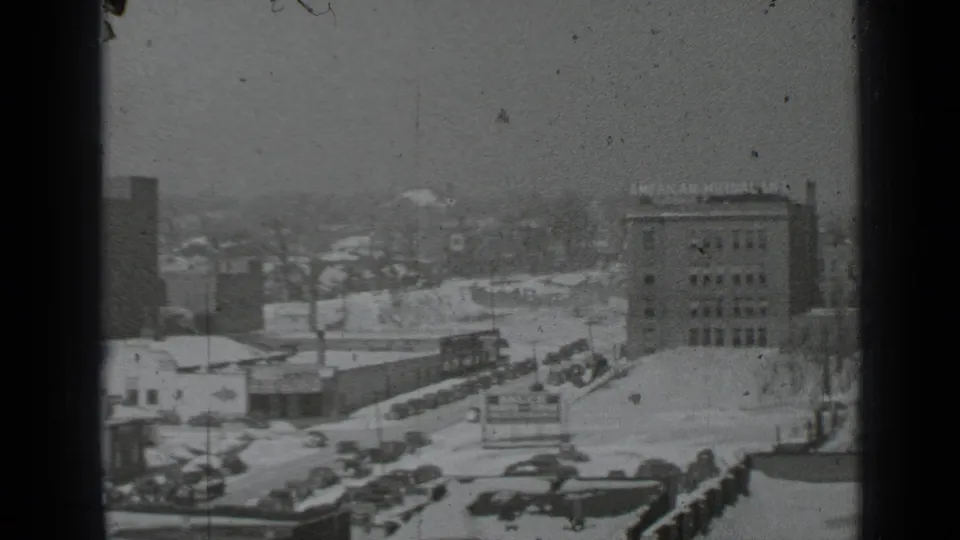 Snowy New York street view from above with parked cars, mid-rise buildings, and an American Museum of Natural History sign visible.