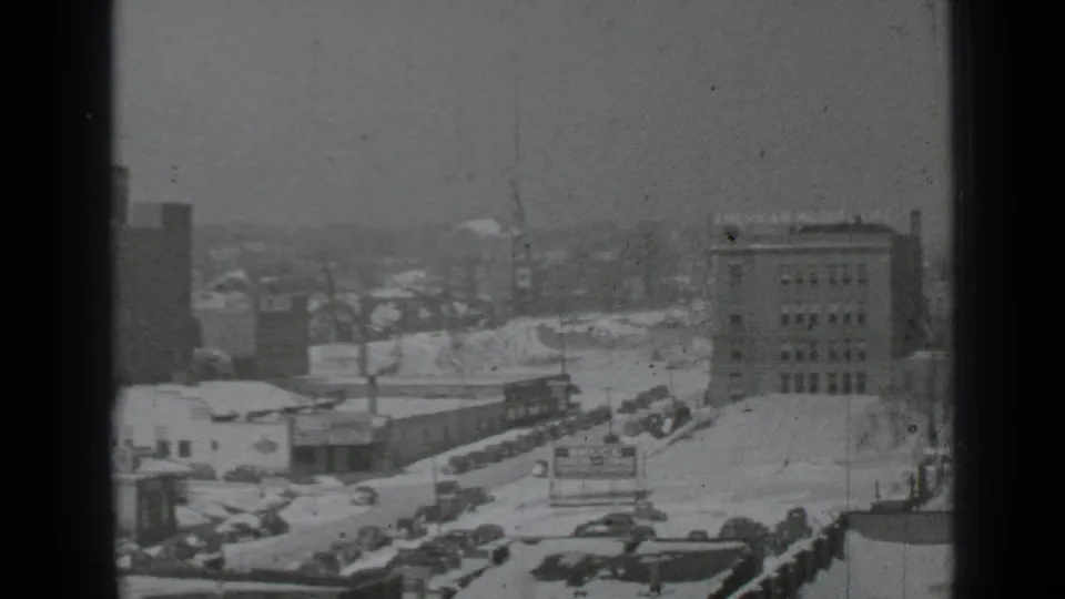 1939 New York winter city view showing a museum sign, snow-covered lots and streets, parked cars, and nearby mid-rise buildings.