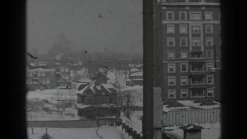 Snow-covered New York cityscape seen from above, with a tall apartment-like building, low rooftops, bare trees, and dark window frames.